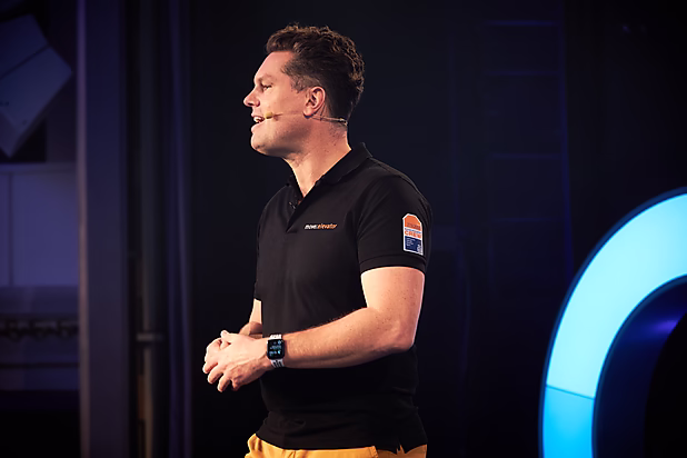 Ein Mann in einem schwarzen Poloshirt spricht leidenschaftlich auf einer Bühne, während er seine Hände bewegt.