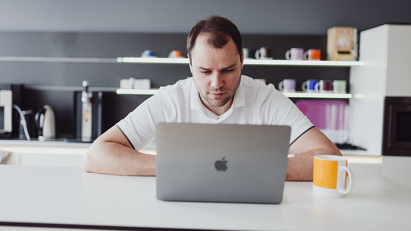 Ein Mann sitzt an einem Tisch und arbeitet konzentriert an einem Laptop in einer modernen Küche. 