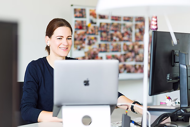 Eine lächelnde Frau sitzt vor einem Laptop, während sie arbeitet. Im Hintergrund sind Fotos an einer Pinnwand zu sehen.