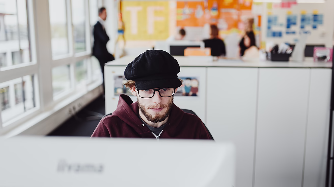 Ein Mann arbeitet an einem Computer in einem modernen Büro. IM Hintergrund sind andere Mitarbeiter in einer BEsprechung an einer Wand mit bunten Notizen.