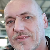 Ein Mann mit kurzen, grauen Haaren und einem Bart schaut in die Kamera. Er hat blaue Augen und trägt einen schwarzen Kragen.