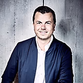 Ein Mann mit kurzen, dunklen Haaren lächelt freundlich, trägt eine dunkelblaue Jacke über einem weißen Hemd. Hintergrund aus Beton.
